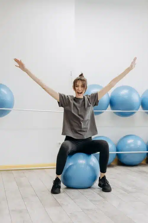 Jeune femme enthousiaste assise sur un gros ballon de gym avec les bras en l'air