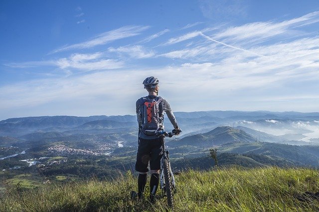 Homme face à un paysage, entre plaine et montagne, tenant son vélo par le guidon.
