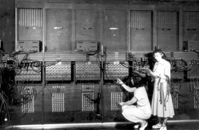 Deux femmes, membres des ENIAC girls, manipulent des câbles sur une machine, qui correspond au premier ordinateur de l'histoire. 