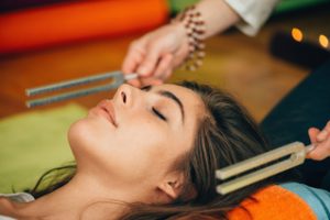 Une femme allongée profite des bienfaits d'un massage sonore. La sonothérapeute fait résonner des diapasons thérapeutiques de chaque côté de sa tête.