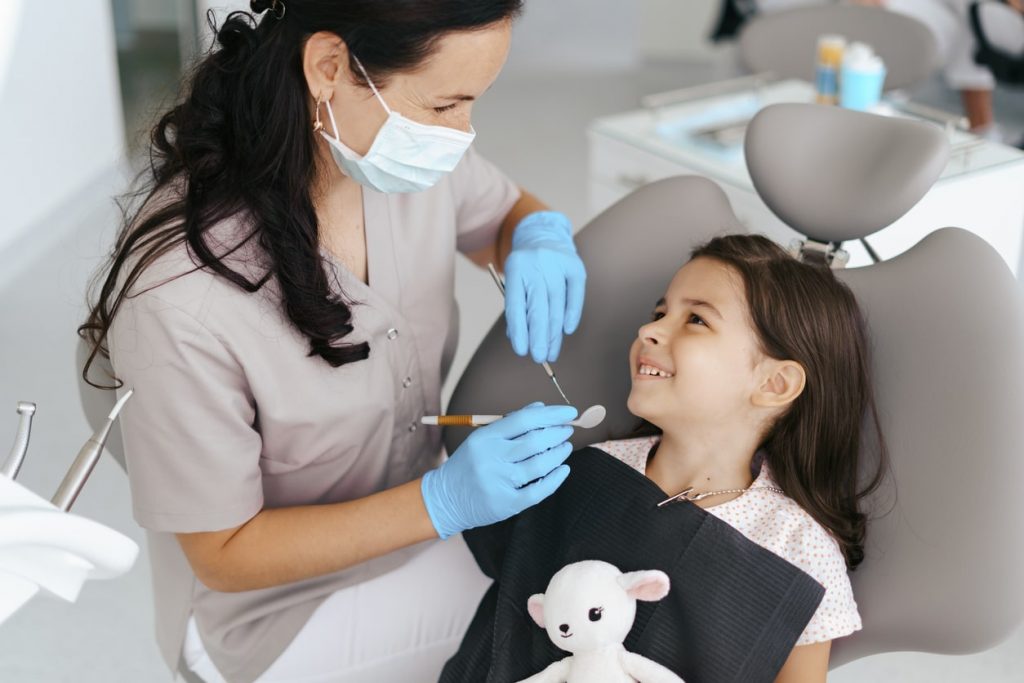 Une petite fille est assise sur le fauteuil de la dentiste. La dentiste approche un miroir et une sonde, la petite fille la regarde en souriant. La praticienne semble avoir instauré une relation de confiance afin d'éviter la peur chez cette enfant. 