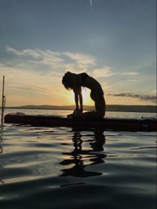 Silhouette d'une personne sur un paddle dans la position du chameau à contre jour devant un coucher de soleil. Cette posture de yoga, exécutée sur une planche, offre de nombreux bénéfices sur la santé.